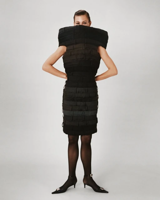 A woman poses in a structured black dress against a white backdrop, accessorized with black tights and pointed heels with jeweled embellishments.
