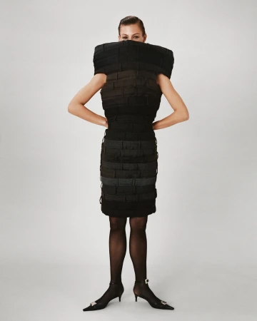 A woman poses in a structured black dress against a white backdrop, accessorized with black tights and pointed heels with jeweled embellishments.