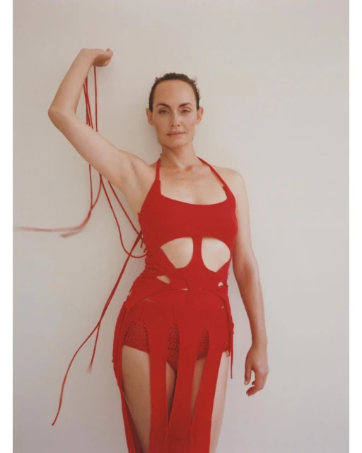 Portrait of a fair-skinned woman in a red halter dress holding red string, posing against a cream-colored wall in a minimalist photo studio.
