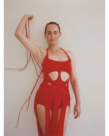 Portrait of a fair-skinned woman in a red halter dress holding red string, posing against a cream-colored wall in a minimalist photo studio.