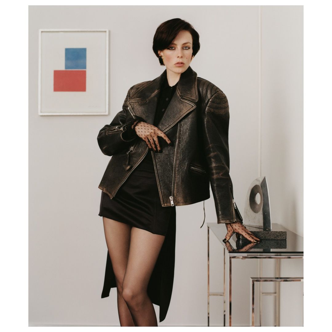 A woman poses in a studio wearing a black mini dress, leather jacket, and dotted mesh gloves, leaning on a glass and metal table.