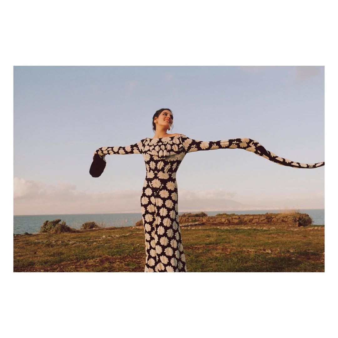 A woman in a long floral dress stands on a grassy cliff overlooking the ocean, arms outstretched under a clear sky at daytime.