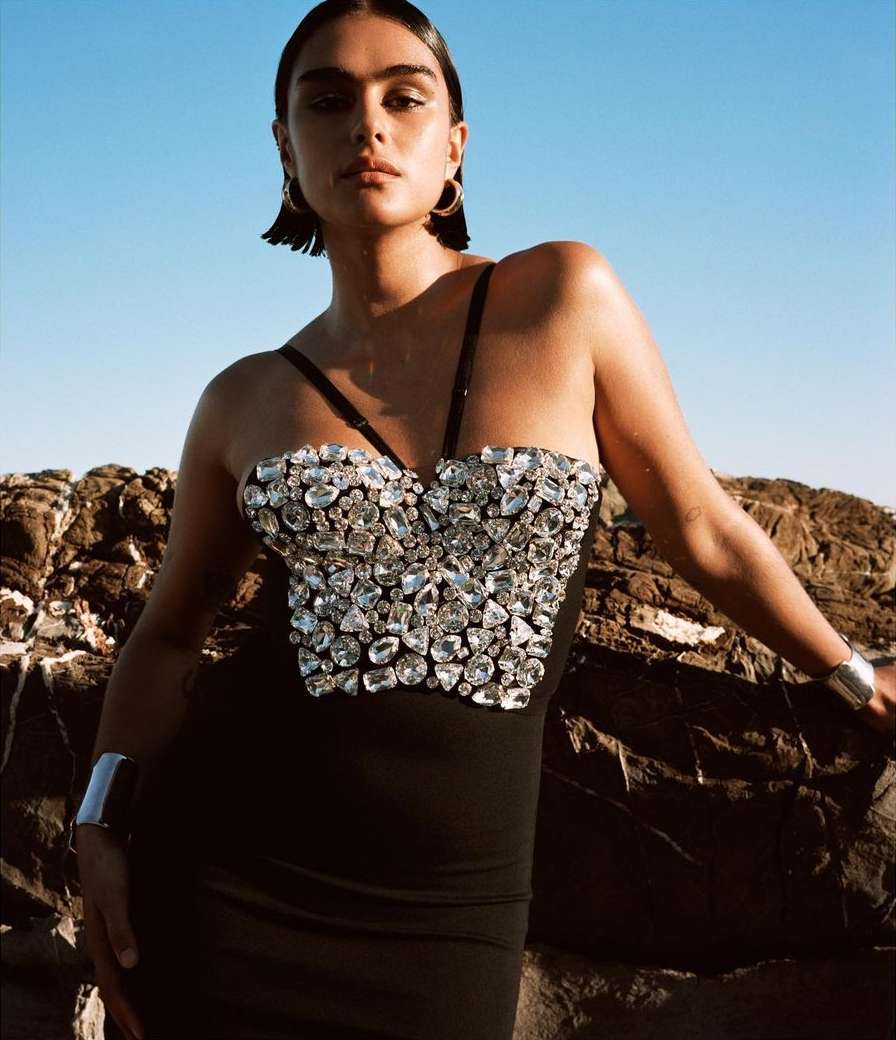 A woman in a black dress with crystal detail poses against a rocky coast under a clear blue sky, showcasing fashion and style.