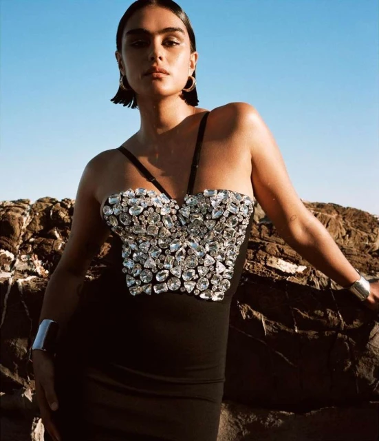 A woman in a black dress with crystal detail poses against a rocky coast under a clear blue sky, showcasing fashion and style.