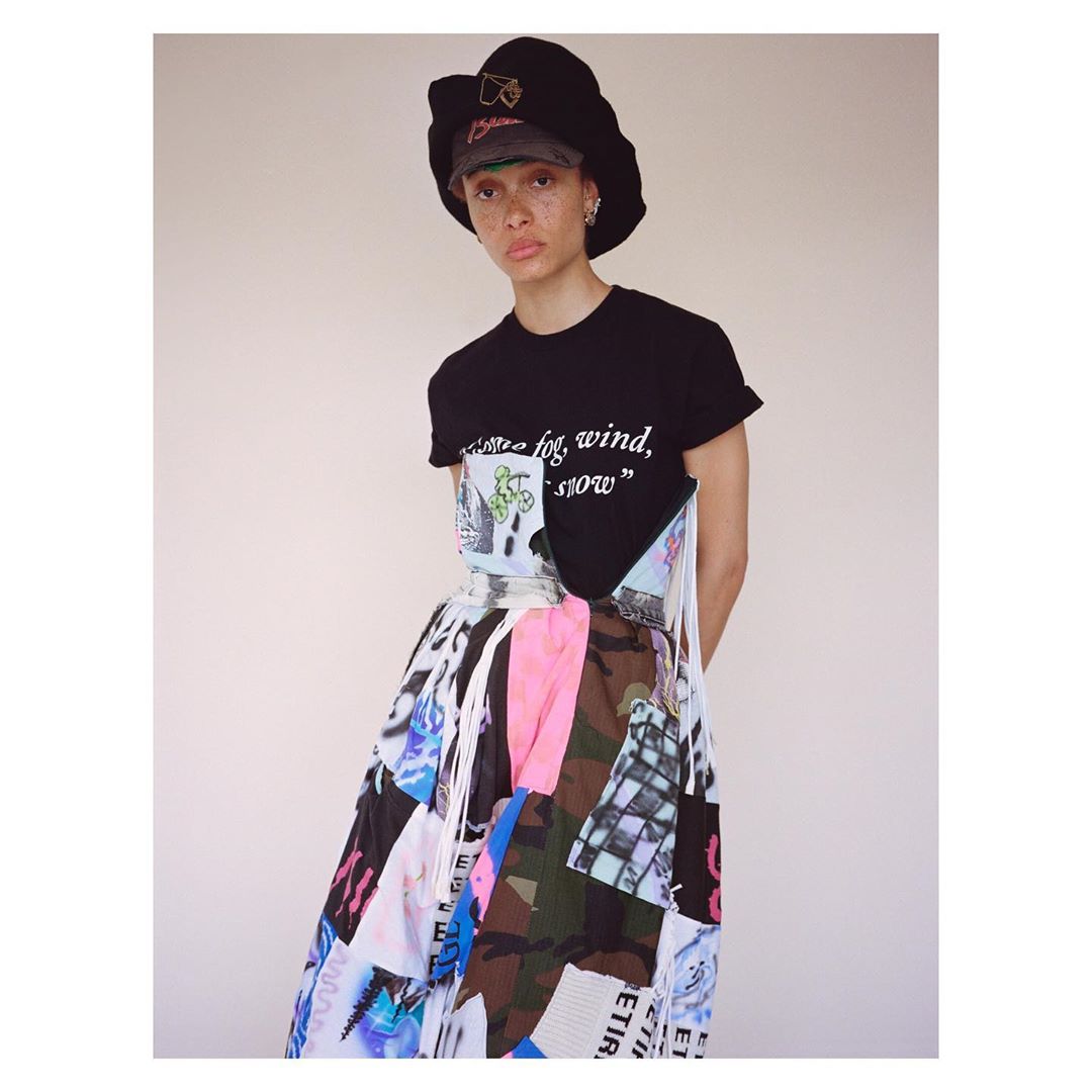 A woman poses in a black t-shirt, colorful patchwork skirt, and bucket hat against a pale background in a fashion-style medium shot.