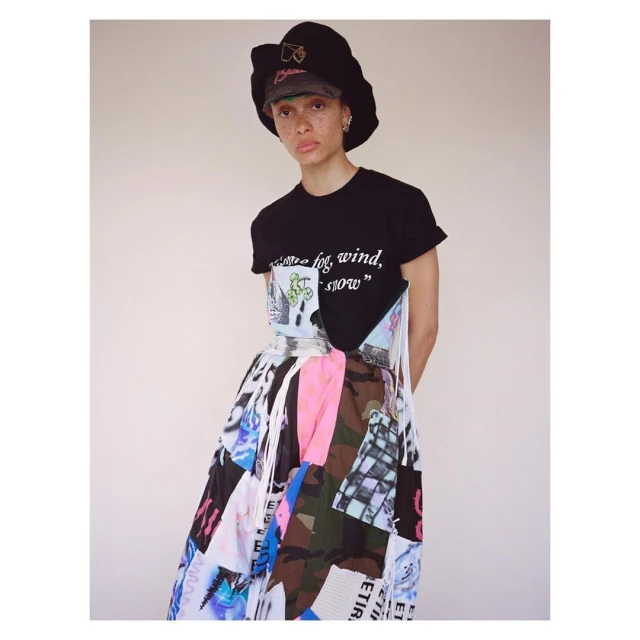 A woman poses in a black t-shirt, colorful patchwork skirt, and bucket hat against a pale background in a fashion-style medium shot.