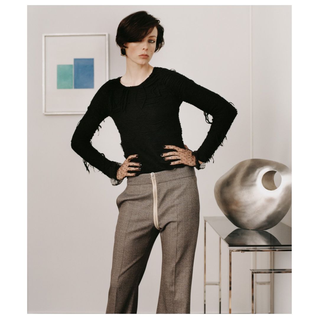 A slender fashion model poses indoors wearing a black fringe sweater and wide-leg grey pants. The setting includes artwork and modern decor.