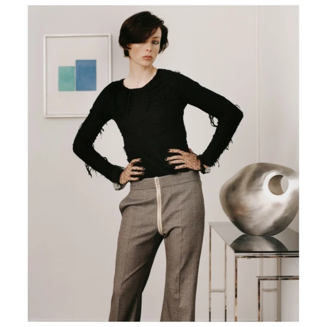 A slender fashion model poses indoors wearing a black fringe sweater and wide-leg grey pants. The setting includes artwork and modern decor.