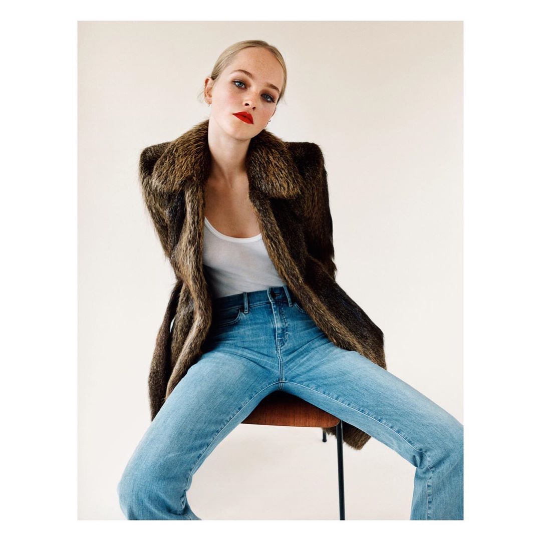 Woman wearing a brown fur collar coat and blue jeans sitting on a chair against a neutral background