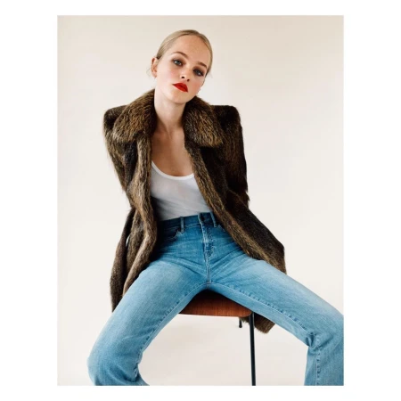 Woman wearing a brown fur collar coat and blue jeans sitting on a chair against a neutral background
