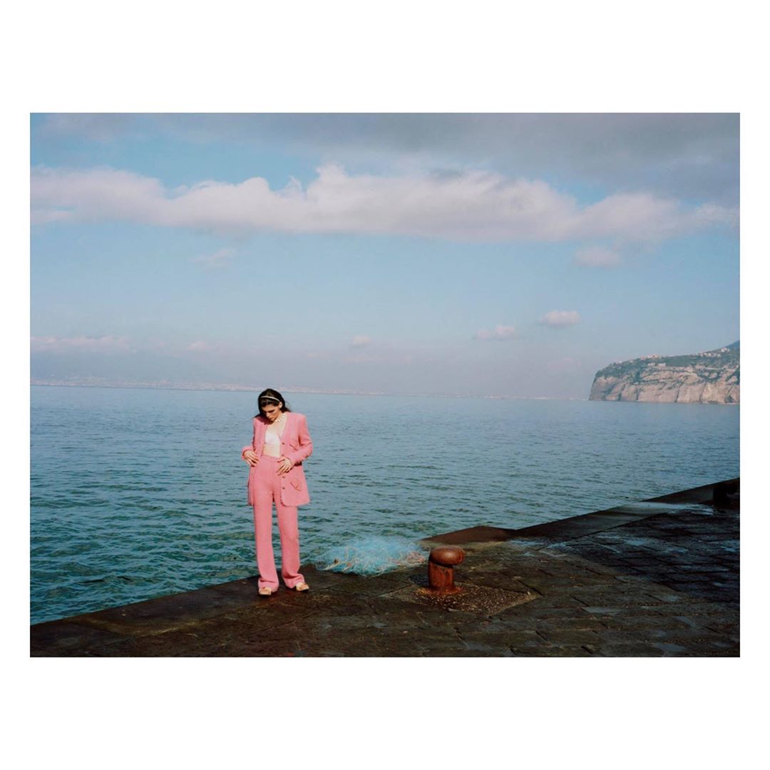 Adult female in a pink suit stands on a stone pier by the ocean, with a cloudy sky and distant mountains in the background.