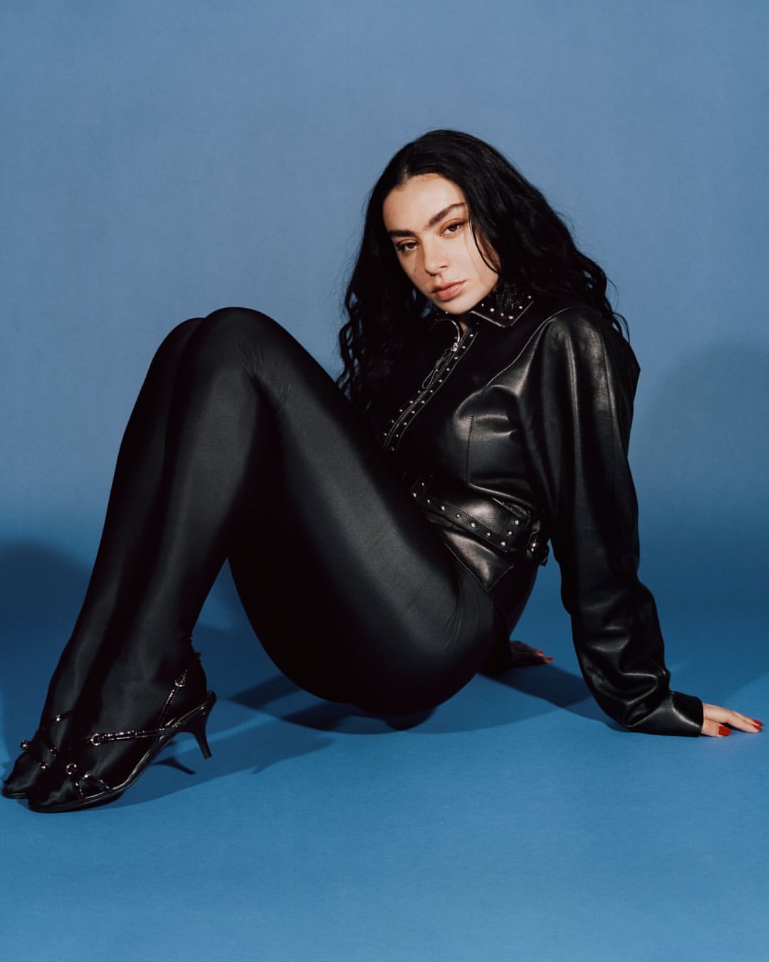 A woman with dark hair poses in a black studded jacket and pants against a blue backdrop, creating a high-fashion editorial image.