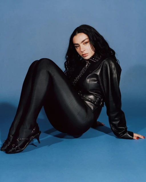 A woman with dark hair poses in a black studded jacket and pants against a blue backdrop, creating a high-fashion editorial image.