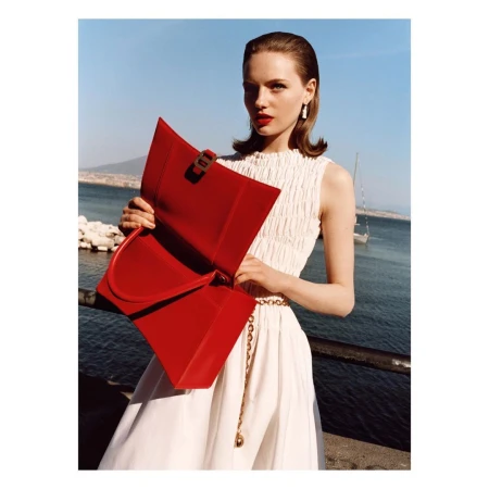 Woman in white dress poses with red leather handbag against coastal backdrop with blue sea and mountain; fashion, accessories, style.