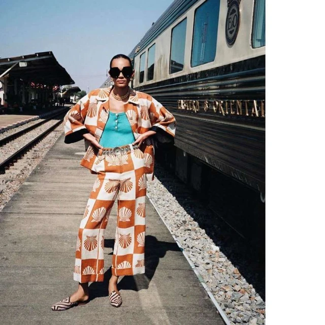 Stylish woman posing on a train platform wearing a patterned co-ord set, sunglasses, and gold jewellery in daytime light.