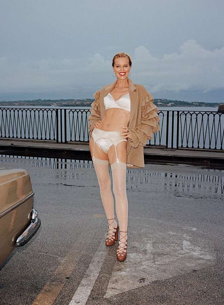 Woman in lingerie and blazer posing on wet city street with car and ocean in background under an overcast sky, looking at camera.