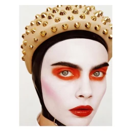 Close-up portrait of a model with white face makeup, orange eye shadow, and a studded beige beret by photographer Angelo Pennetta.