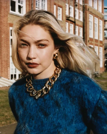Gigi Hadid wearing a blue textured sweater and chunky gold chain necklace outdoors against a brick building