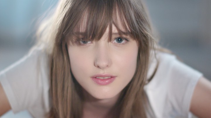 Close-up portrait of a young adult woman with fair skin, bangs, blue eyes, and a neutral expression, wearing a white top against a soft background.