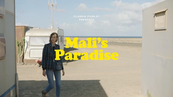 A woman in a blazer and jeans walks on a sandy beach near a vintage trailer. The ocean is visible in the background under a bright sky.