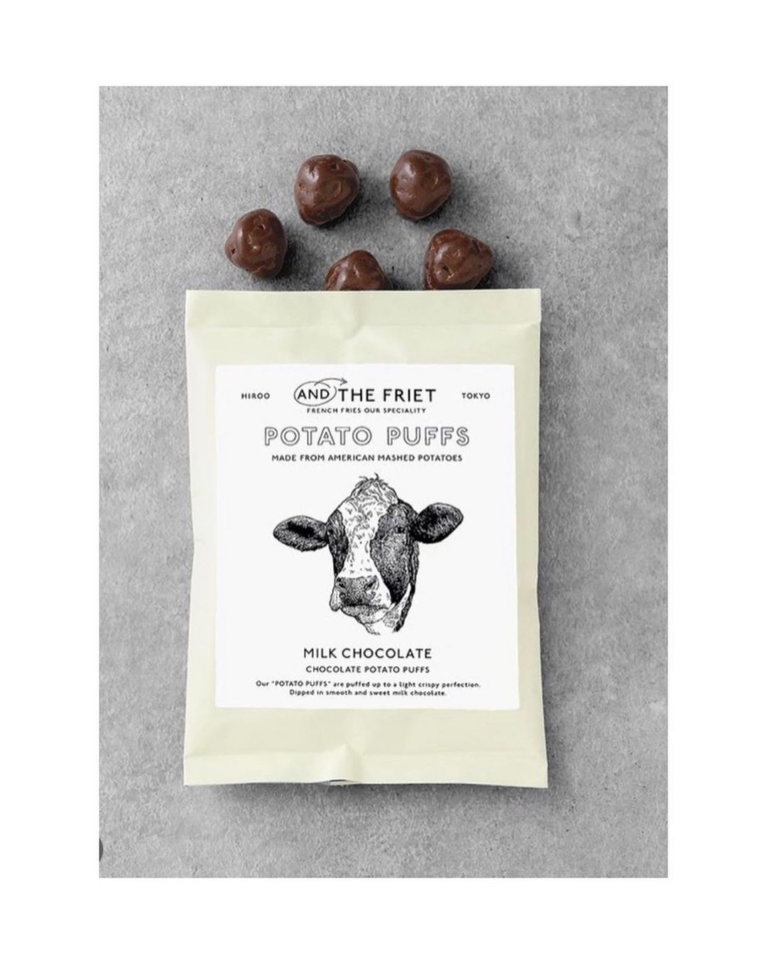 Potato puffs in a bag with milk chocolate, featuring a cow illustration, presented on a gray surface with several chocolate pieces.