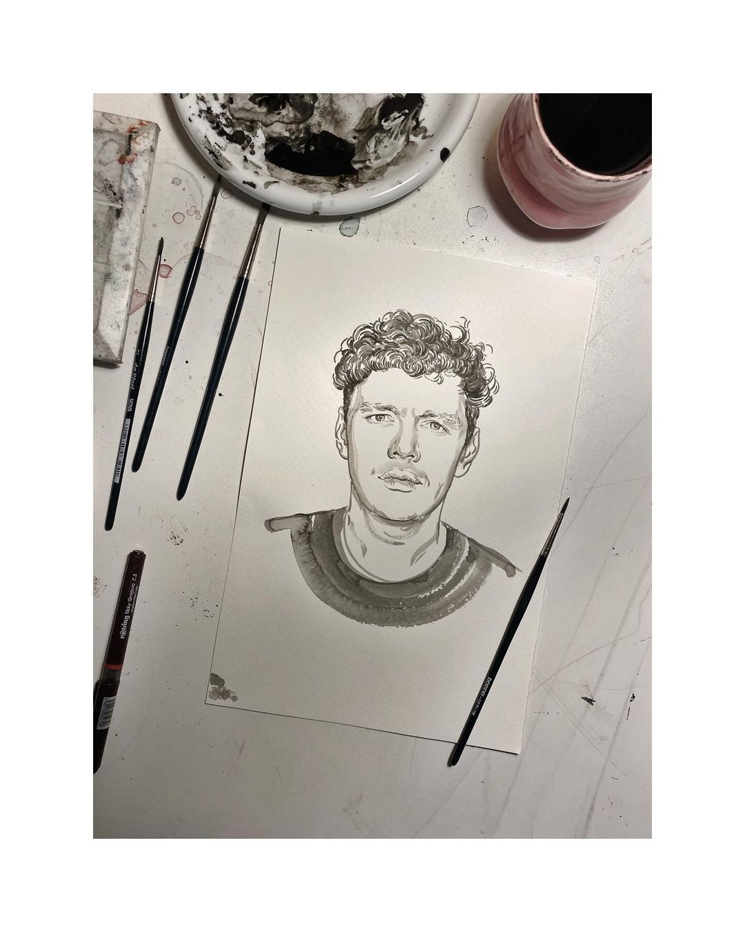 Monochrome ink portrait of a young adult with curly hair, painted on white paper, surrounded by paintbrushes and an ink palette.