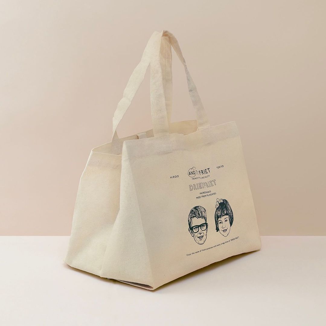 Cream-colored canvas tote bag featuring illustrations of two people and the brand logo in a studio setting. Sustainable fashion.