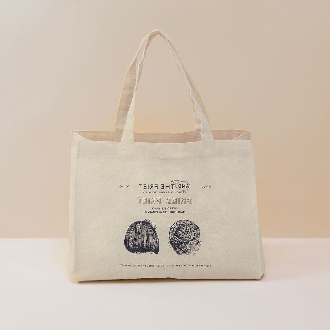 Beige tote bag with graphic print of two hair styles and the text “Dried Fries.” Modern style on a light beige background in studio.