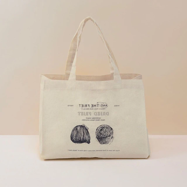 Beige tote bag with graphic print of two hair styles and the text “Dried Fries.”  Modern style on a light beige background in studio.