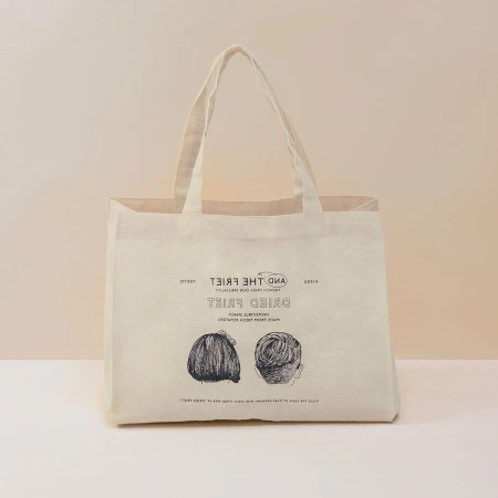 Beige tote bag with graphic print of two hair styles and the text “Dried Fries.”  Modern style on a light beige background in studio.