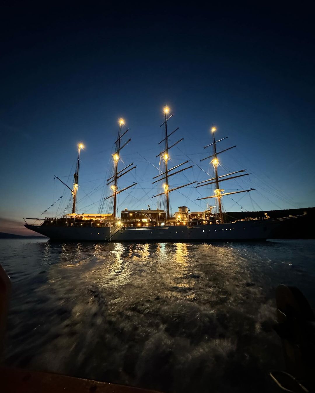 Illuminated tall ship with multiple masts sails on calm water in the evening. The sky is clear with a blue and black gradient.