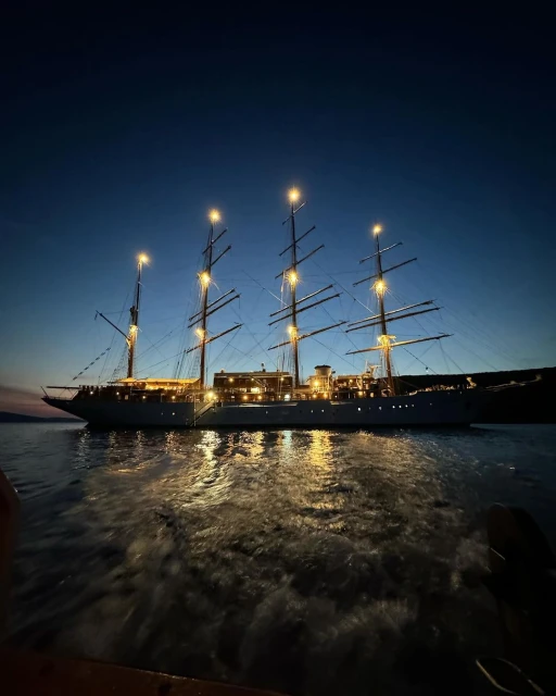 Illuminated tall ship with multiple masts sails on calm water in the evening. The sky is clear with a blue and black gradient.