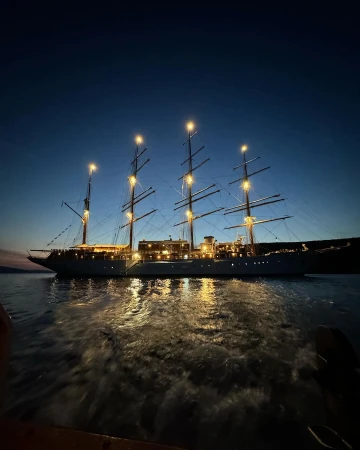 Illuminated tall ship with multiple masts sails on calm water in the evening. The sky is clear with a blue and black gradient.