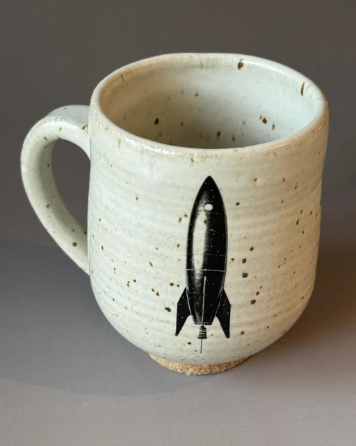 Ceramic mug with a cylindrical shape and a handle, featuring a black rocket graphic, set against a neutral grey backdrop.