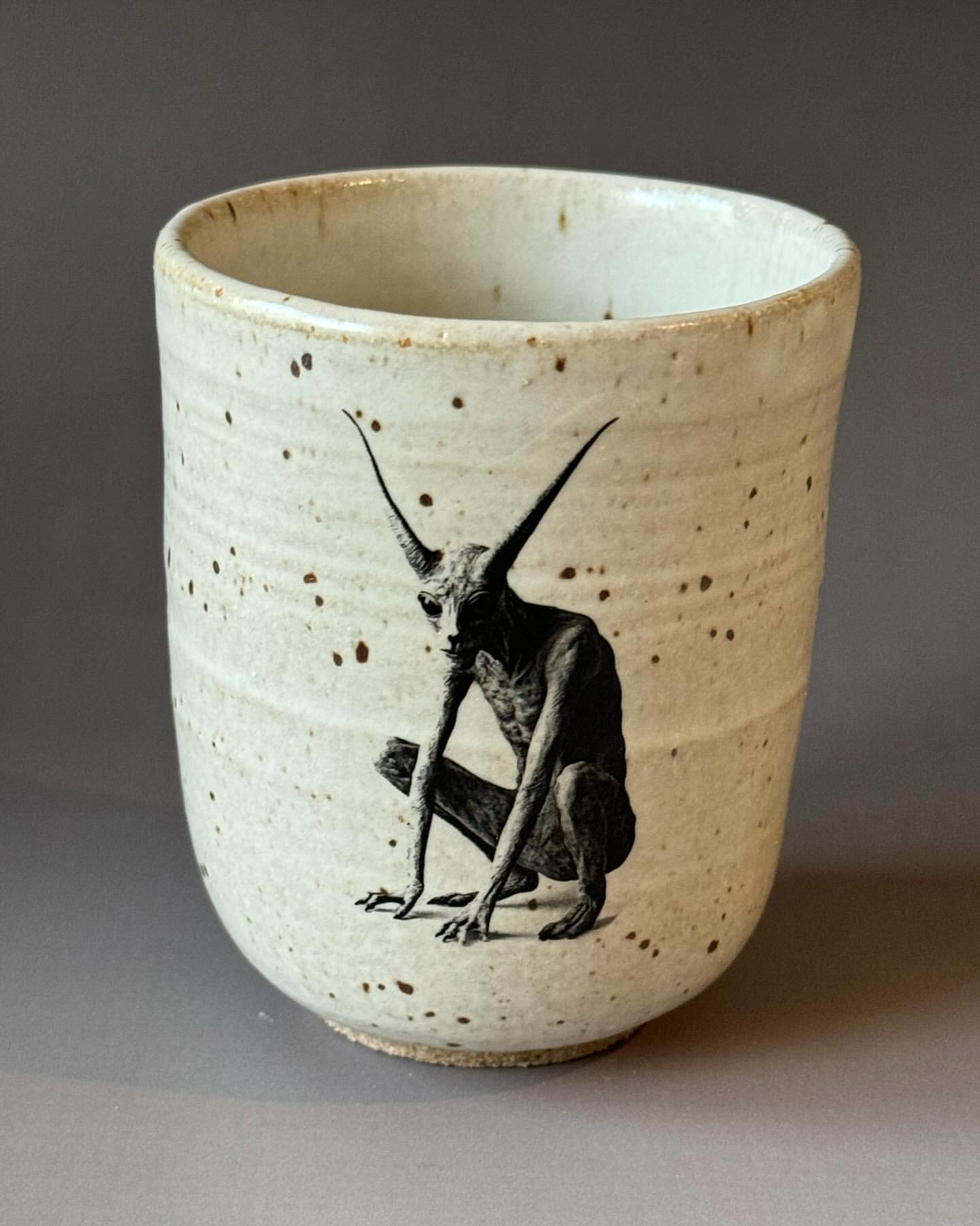 Close-up studio shot of ceramic cup featuring an illustration of a horned, humanoid creature, set against a neutral gray background.