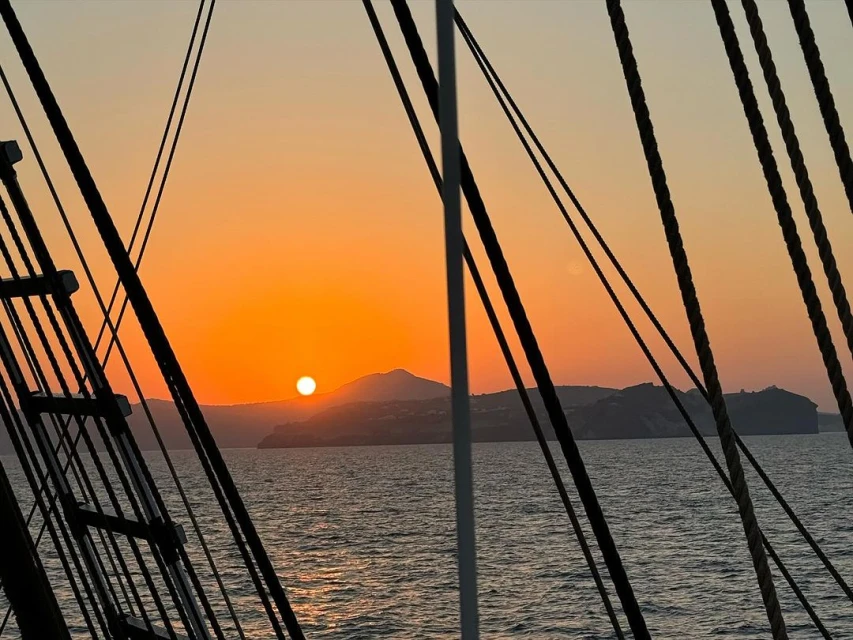 Sunset over the ocean with silhouetted islands in the background, framed by the rigging of a boat in the foreground.