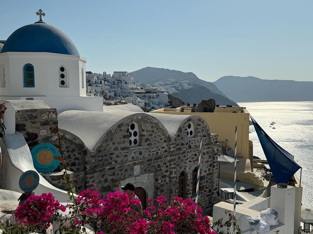 Scenic view of a Greek island with a traditional white church with blue dome, pink flowers, and the Aegean sea in the background.