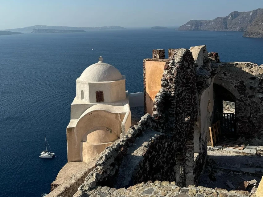 Santorini, Greece vista showcasing a white domed church alongside stone ruins overlooking the Aegean Sea, with a catamaran sailboat visible in the distance.