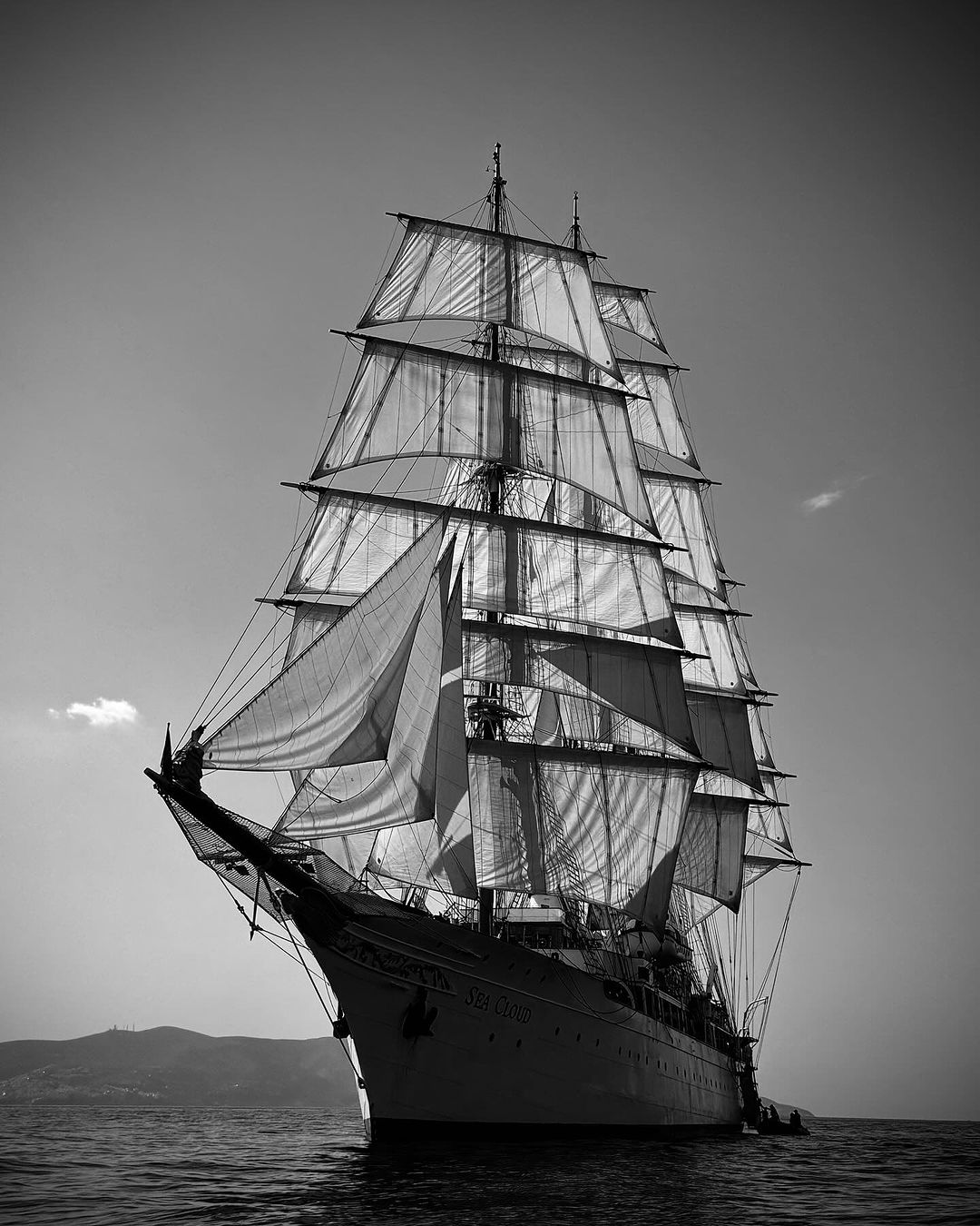 Black and white shot of a tall ship sailing on the open ocean, with billowing sails and dark water in the foreground.