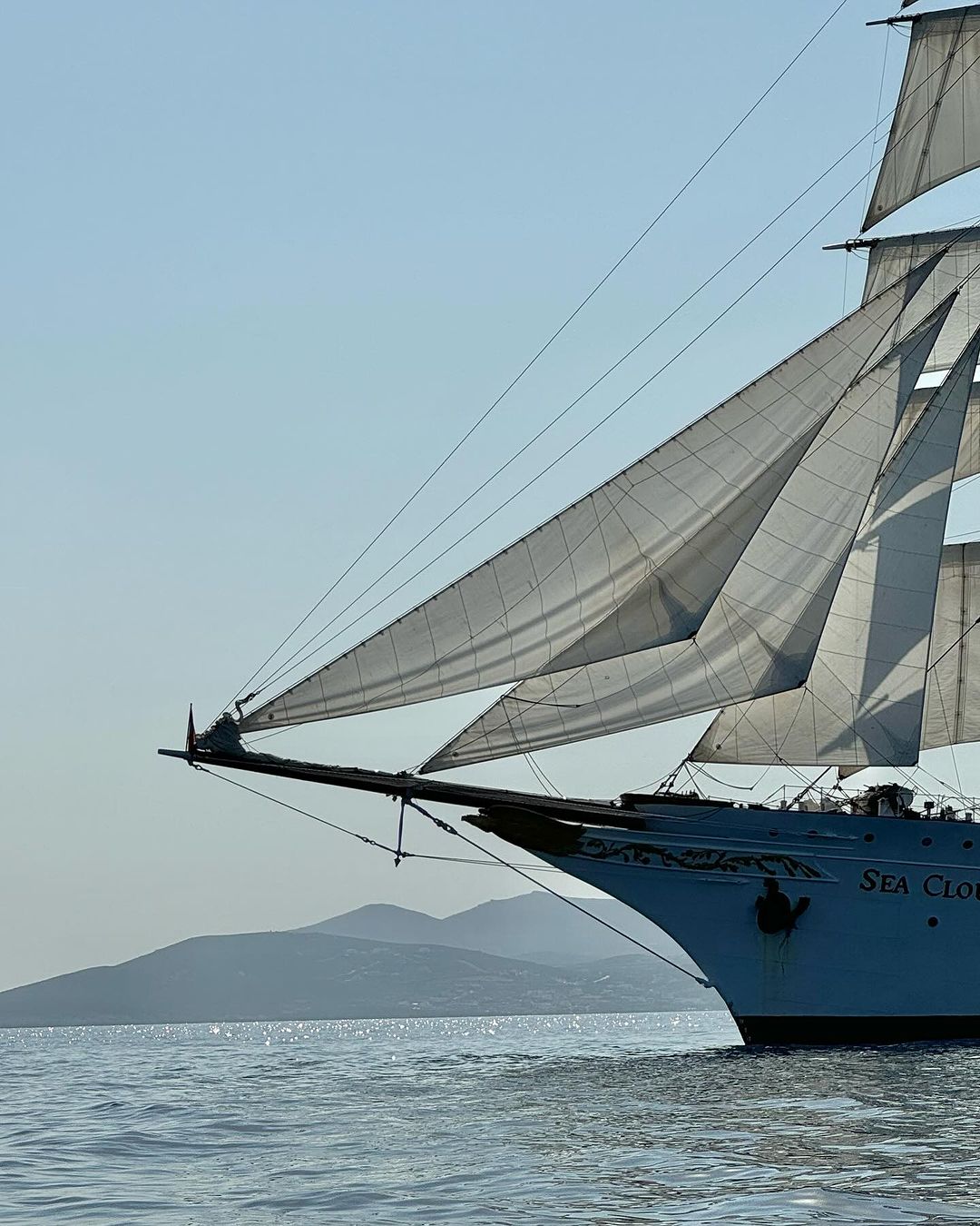 Sailing ship with white sails on a calm sea under a clear sky, with a hint of land in the background, creating a serene nautical scene.