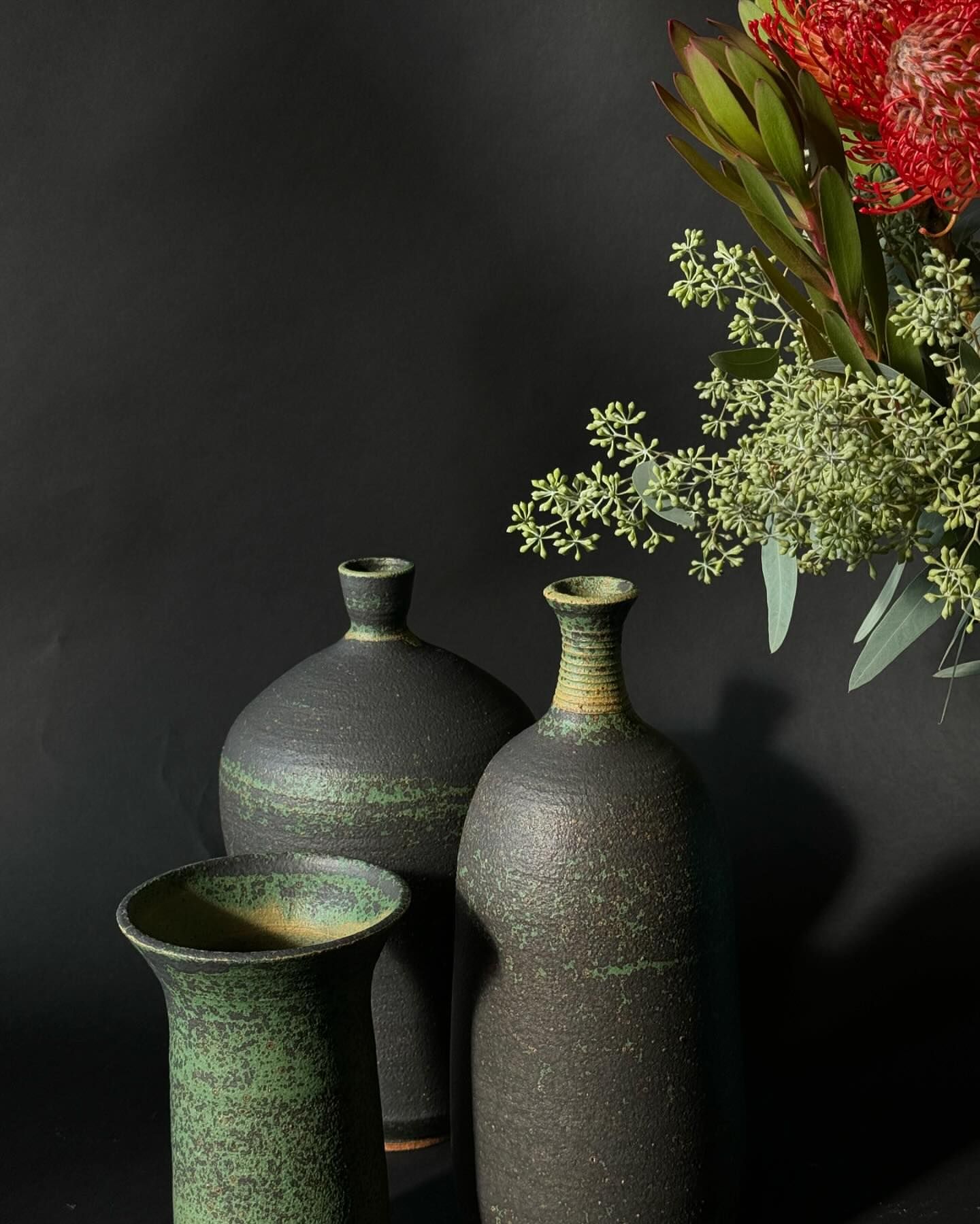 Three handmade vases of different sizes, two black and one green, are set against a dark backdrop with floral elements on the right.