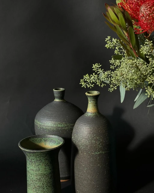 Three handmade vases of different sizes, two black and one green, are set against a dark backdrop with floral elements on the right.