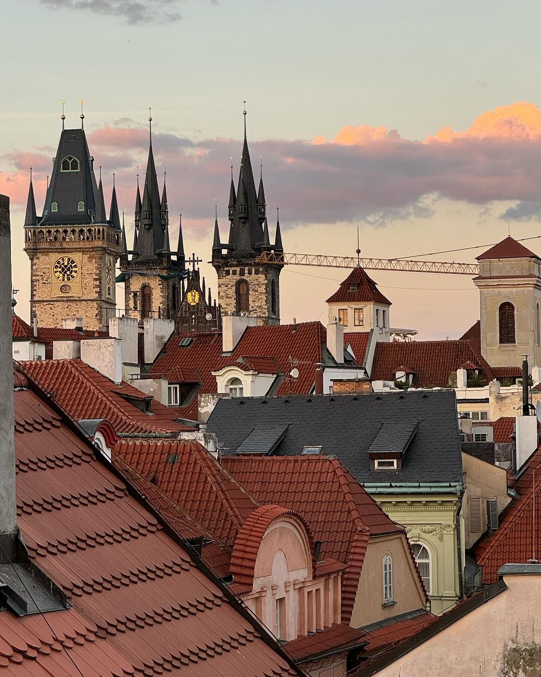 View of Prague rooftops and architecture with spires against a colorful sunset sky. Buildings feature red tiles and ornate clock towers.