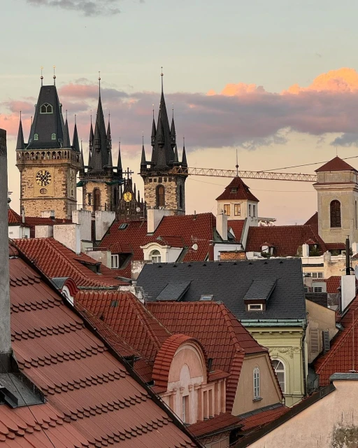 View of Prague rooftops and architecture with spires against a colorful sunset sky. Buildings feature red tiles and ornate clock towers.