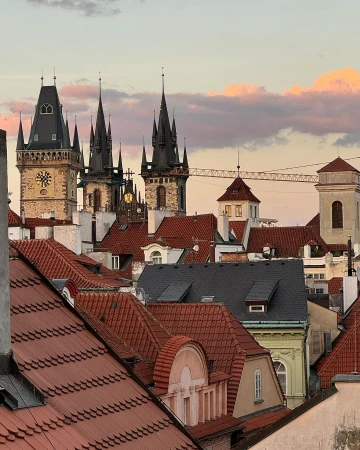 View of Prague rooftops and architecture with spires against a colorful sunset sky. Buildings feature red tiles and ornate clock towers.