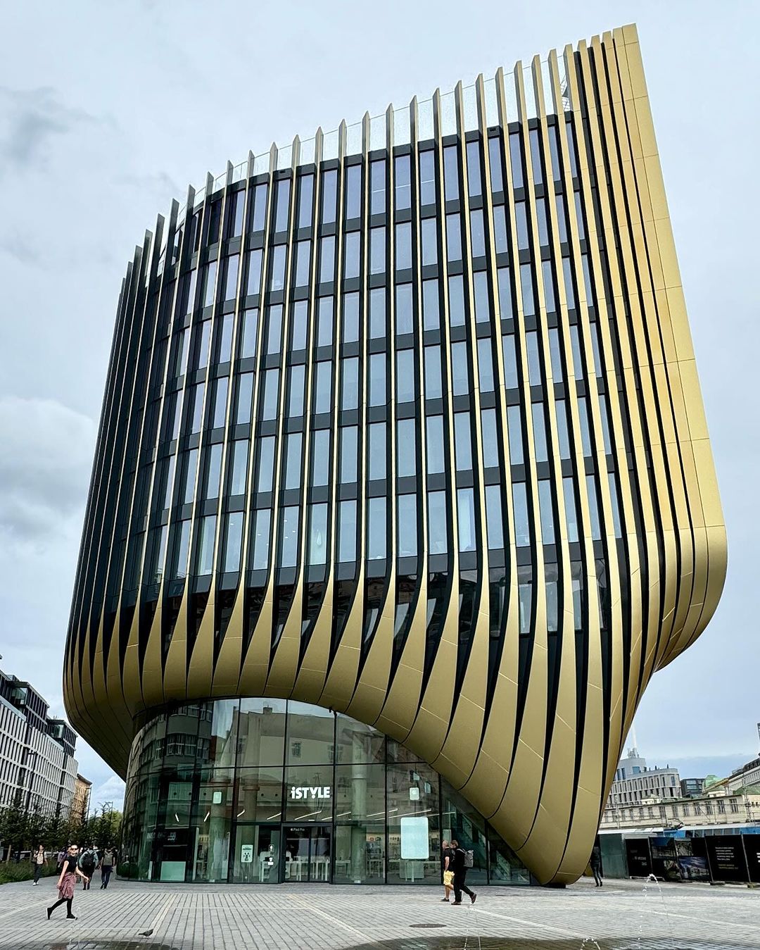 Exterior of the modern iSTYLE building featuring curved gold cladding, many windows, and urban setting on a cloudy day.