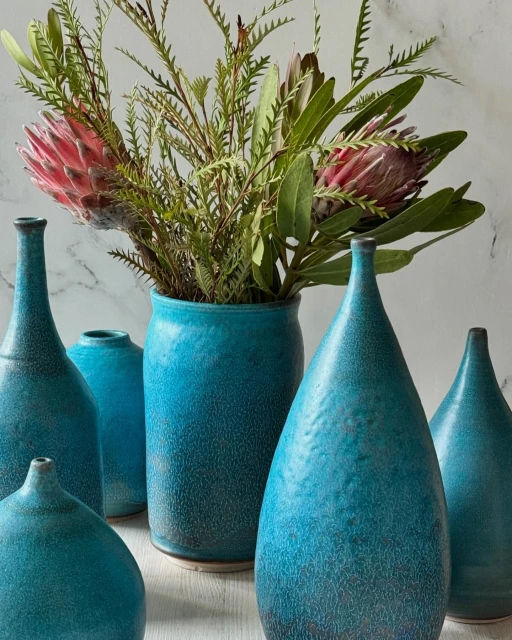 Still life of blue ceramic vases and flower arrangement against a textured white backdrop. Features various vase shapes and floral elements.