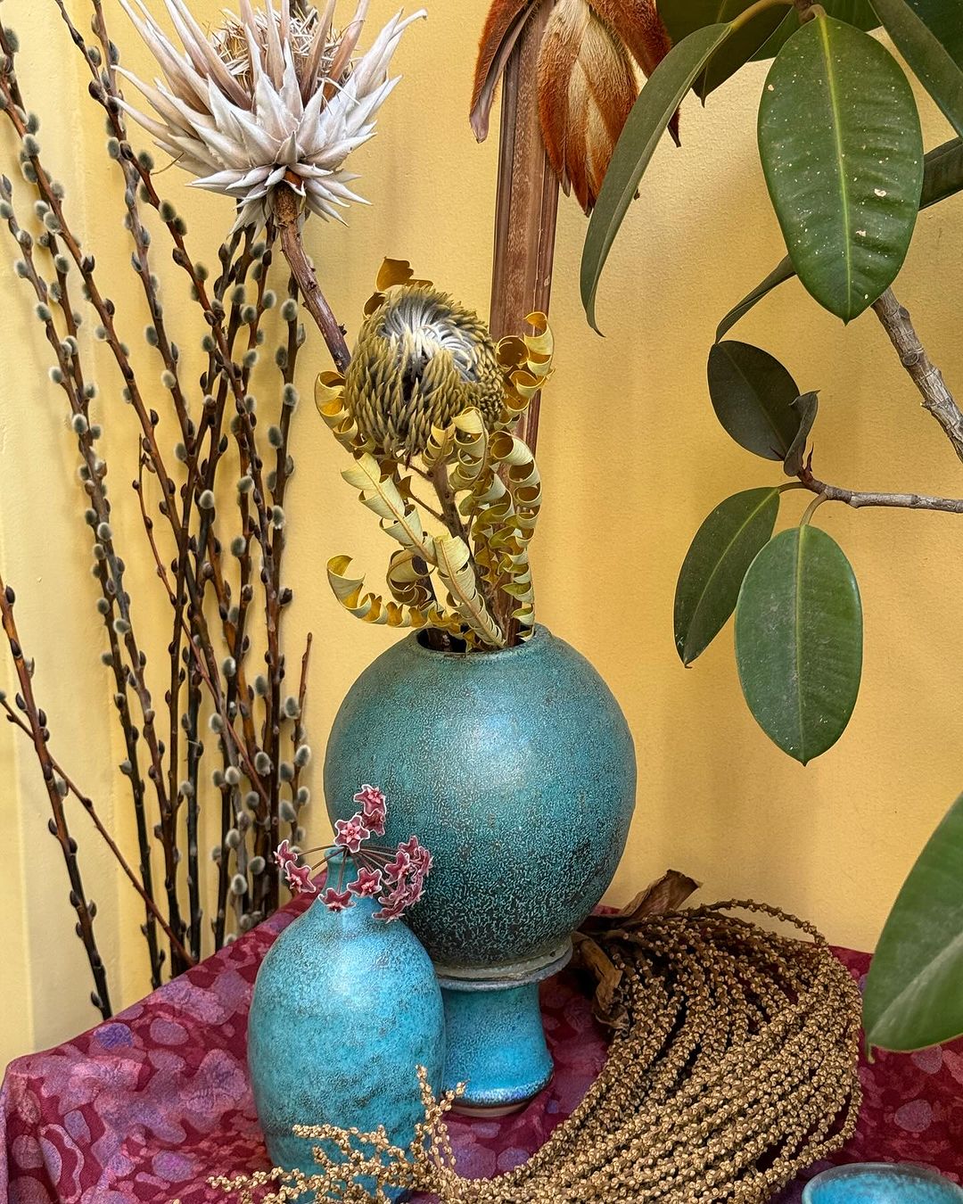 Arrangement of dried flowers and plants in round turquoise vases on a patterned surface against a yellow wall in a close-up shot.