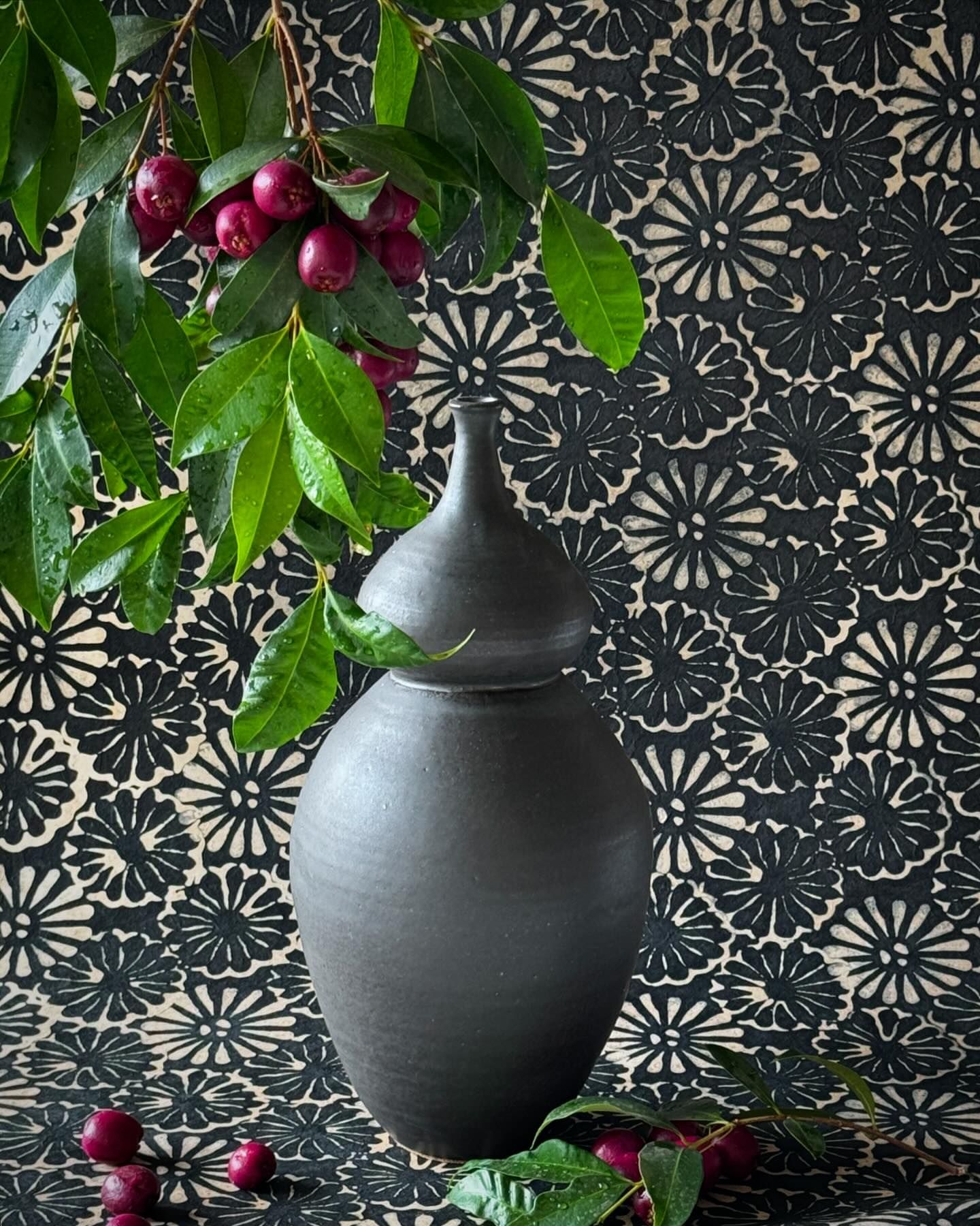 Still life photograph of a black ceramic vase with a lid, decorated with green leaves and red berries, set against a floral backdrop.