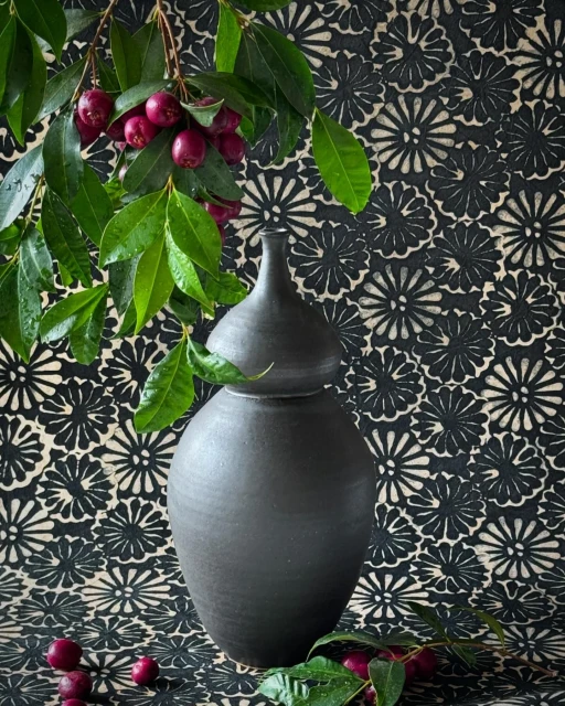 Still life photograph of a black ceramic vase with a lid, decorated with green leaves and red berries, set against a floral backdrop.